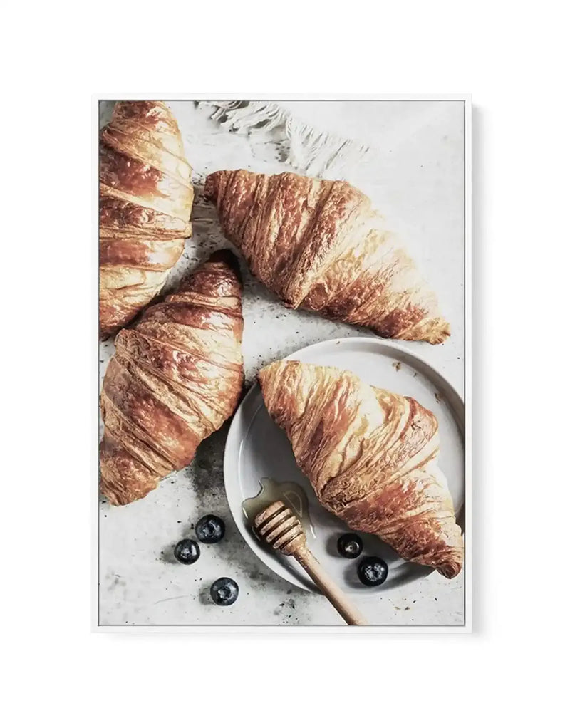 Framed canvas art print featuring a flat-lay food photography composition. Three golden-brown croissants with layered, flaky textures are arranged on a light gray speckled surface alongside a white plate containing a honey dipper and scattered fresh blueberries. Warm tan and rust tones dominate the image, creating an inviting, artisanal bakery aesthetic. The canvas is mounted in a white frame with white border.
