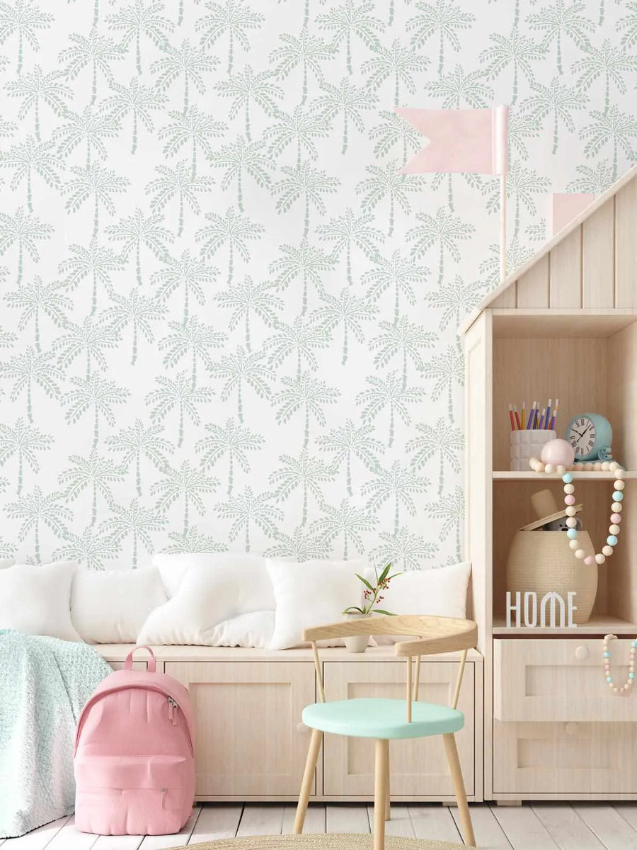 Lifestyle shot of a child's room featuring Cruisey Palms wallpaper in mint. The wallpaper has a repeating pattern of hand-drawn palm trees in a soft mint green on a white background, creating a playful tropical feel. The room includes a built-in wooden bench with white cushions, a light wooden chair with a mint seat, a pink backpack, and a wooden shelving unit with various decorative items.