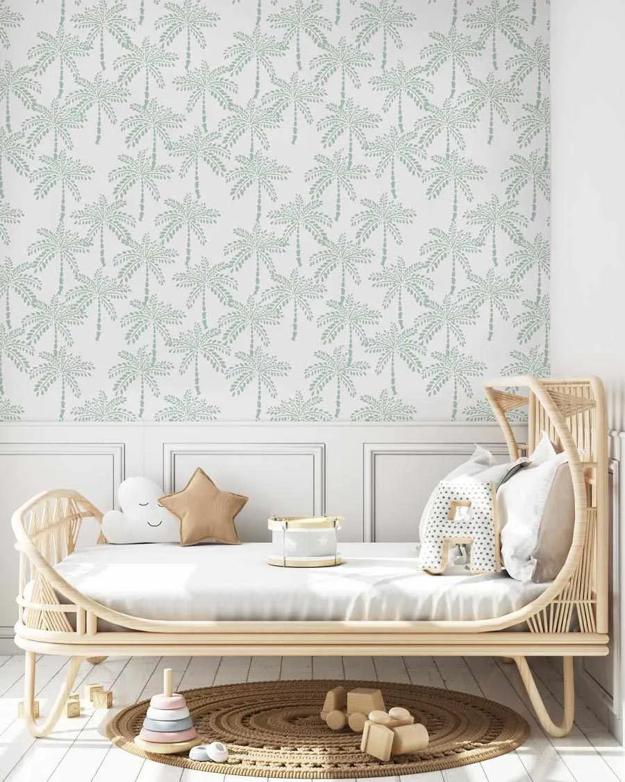 Nursery:
"Peel and stick wallpaper featuring a repeating pattern of hand-painted palm trees in a muted mint green on a soft white background, displayed in a child's nursery. The wallpaper covers the upper half of the wall above white wainscoting. A light wood rattan crib with white bedding is centered against the wall, adorned with a white cloud pillow, a tan star pillow, and a small white and grey toy drum. On the light wood floor, a round woven rug holds wooden toys and a pastel stacking toy."