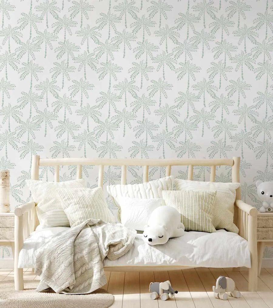 Lifestyle shot of a child's room featuring wallpaper with a repeating pattern of hand-drawn palm trees in a muted mint green on a white background. A light wood daybed with white bedding and several textured cream and white pillows sits against the wall. A white polar bear plush toy and two small elephant toys are on the floor.