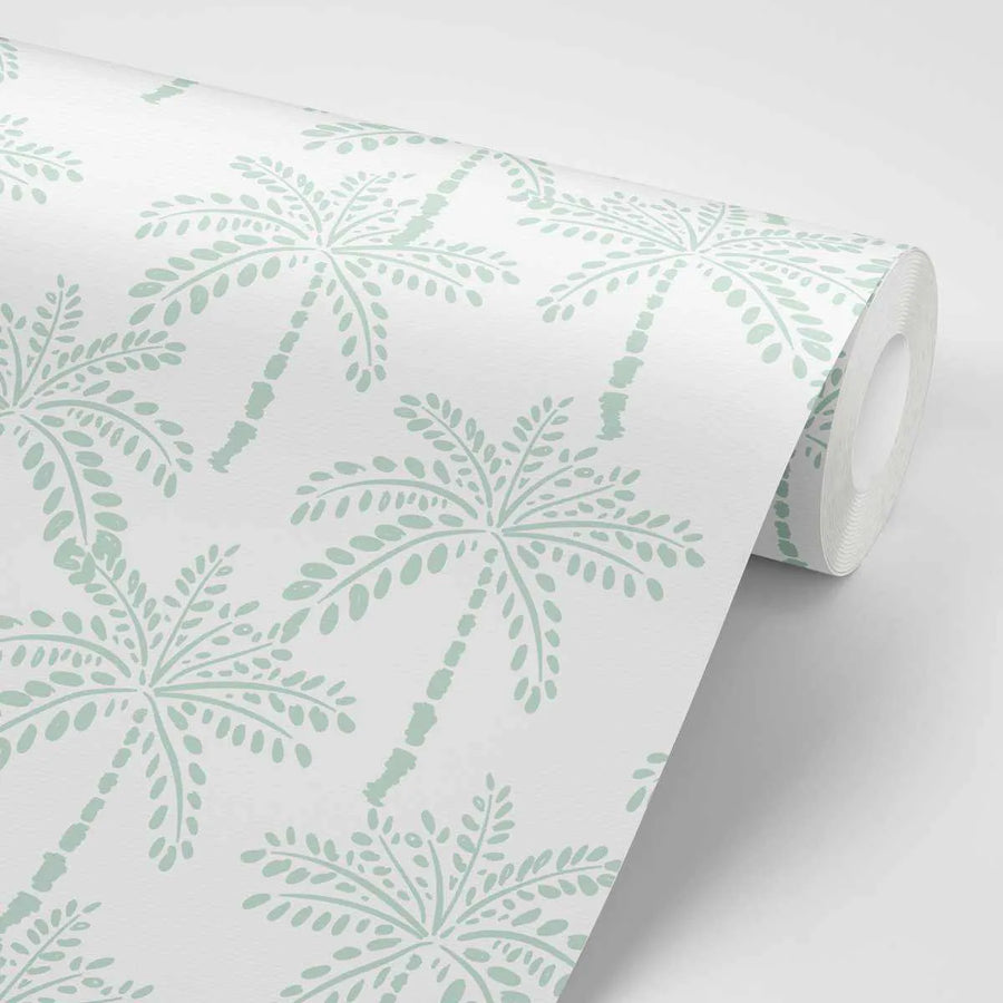 Peel and stick wallpaper featuring a repeating pattern of hand-drawn palm trees in a muted mint green on a clean white background. The design has a slightly distressed, block-print texture, with visible variations in the mint green colour. The wallpaper is shown rolled, with the pattern visible on the unrolled portion.
