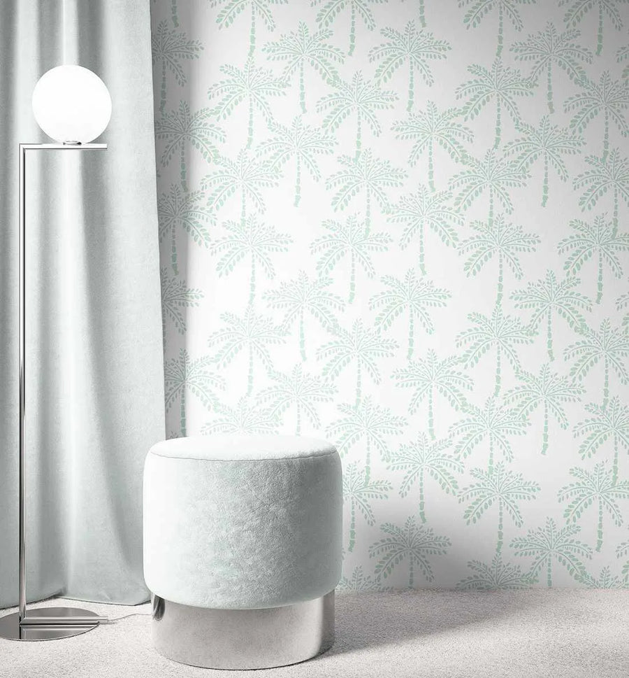 Lifestyle shot of a room featuring Cruisey Palms | Mint Wallpaper. The wallpaper has a repeating pattern of hand-drawn palm trees in a soft mint green against a crisp white background, creating a fresh, tropical feel. The room includes a light grey curtain on the left, a modern floor lamp with a spherical white light, and a round, light grey velvet ottoman with a silver base on a light grey carpet.
