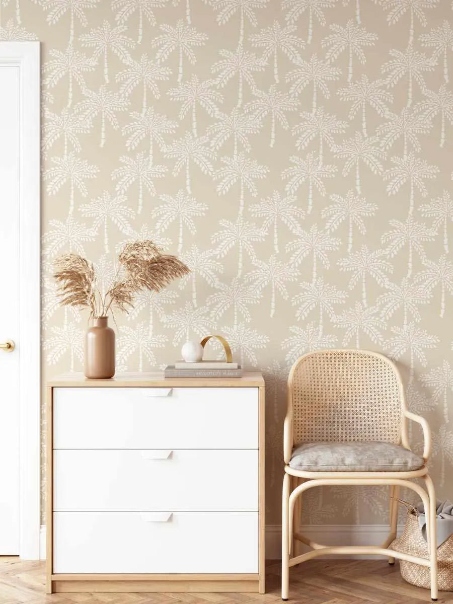 Lifestyle shot of a room featuring Cruisey Palms in Biscuit wallpaper. The wallpaper has a repeating pattern of hand-drawn palm trees in off-white against a warm biscuit beige background. The room includes a white three-drawer dresser with a light wood top, a light wood and cane chair with a grey cushion, and a vase of dried pampas grass.