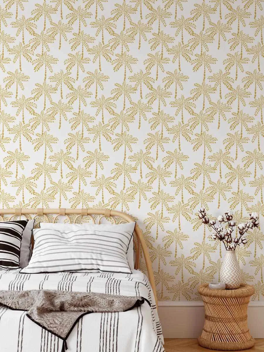 Peel and stick wallpaper featuring a repeating pattern of hand-drawn palm trees in a muted bronze-gold colour on a crisp white background, displayed in a bedroom setting. The wallpaper covers the wall behind a light wood rattan headboard and a bed dressed in white and black striped bedding. To the right, a woven rattan side table holds a white vase with a chevron pattern, filled with cotton branches.