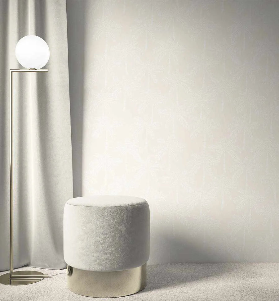 Lifestyle shot of a room featuring Cruisey Palms in Sand Wallpaper. The wallpaper has a subtle, repeating pattern of palm trees in a slightly lighter shade of sand on a warm, light beige background, creating a textured and serene feel. The room includes a tall, slender brass floor lamp with a spherical white globe light, positioned to the left against a floor-length, light beige velvet curtain. In the foreground, a round, light grey velvet ottoman with a brass base sits on a light beige carpet.