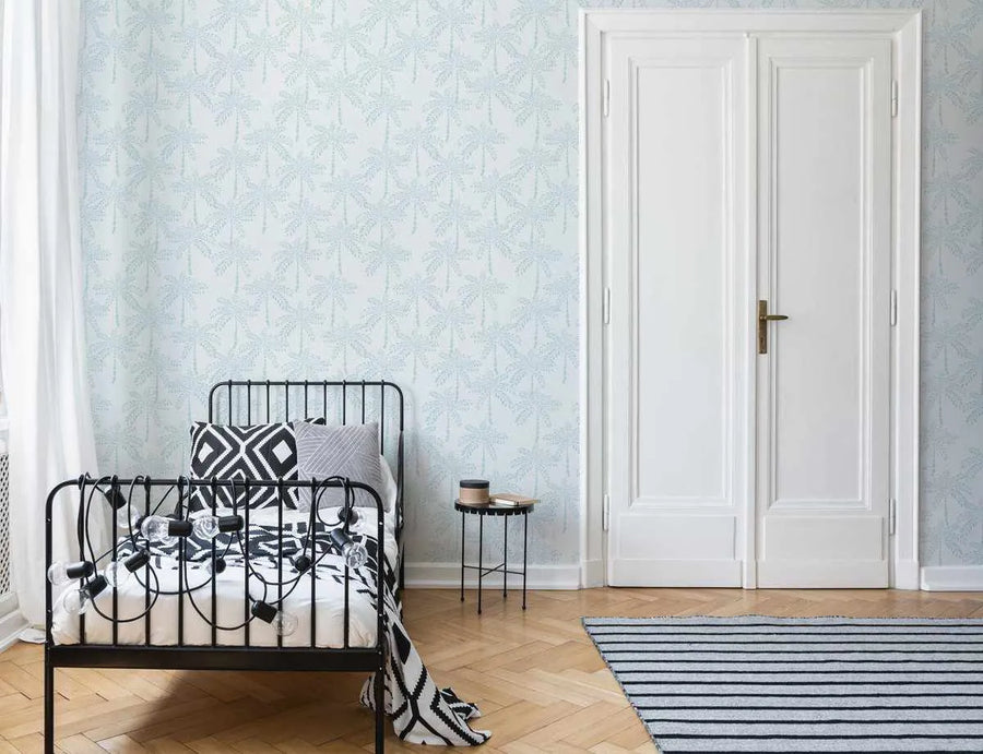 Lifestyle shot of a bedroom featuring Cruisey Palms in Sky Blue Wallpaper. The wallpaper has a repeating pattern of hand-painted palm trees in a soft sky blue against a light cream background, giving a tranquil, tropical feel. The room includes a black metal bed frame with white and black patterned bedding, a small black side table, and a white double door. A striped rug in black and white is visible on the light wood floor.