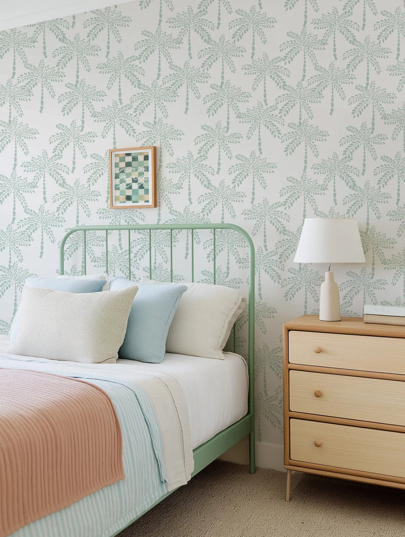 Lifestyle shot of a bedroom featuring peel and stick wallpaper with a repeating pattern of hand-painted palm trees in a muted mint green on a soft white background. The wallpaper has a loose, organic quality with visible brushstroke texture. The room includes a light green metal bed frame with white, light blue, and textured cream pillows, a light pink ribbed throw blanket, and a light wood bedside table with a cream lamp and stacked books.