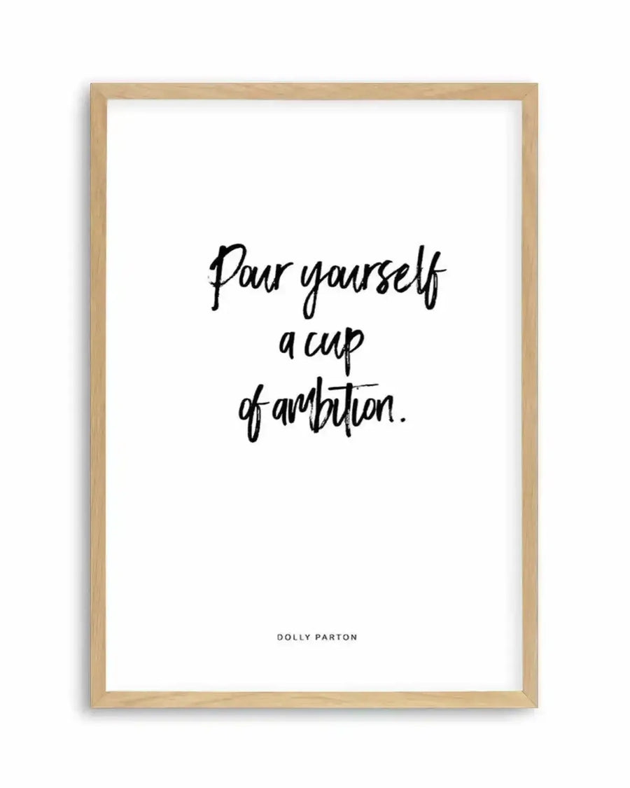 Art print featuring the quote "Pour yourself a cup of ambition." in a bold, handwritten-style black font on a crisp white background, displayed in a natural oak frame with white border. The quote is attributed to Dolly Parton in smaller, sans-serif black text at the bottom.