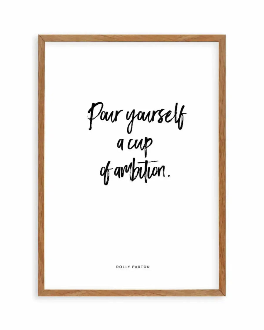 Typography art print featuring the quote "Pour yourself a cup of ambition." in a handwritten-style black script on a white background, printed on fine art paper and displayed in a natural oak frame with white border.