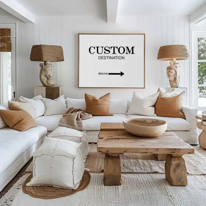 Large framed art print displayed above a white sectional sofa in a bright, coastal-inspired living room. The print is a white signpost-style design with black text reading "CUSTOM DESTINATION" and "804 MI" with a right-pointing arrow. The print has a natural wood frame and white border. The room features a white shiplap wall, light wood and woven textures, and accent pillows in cream and warm tan.