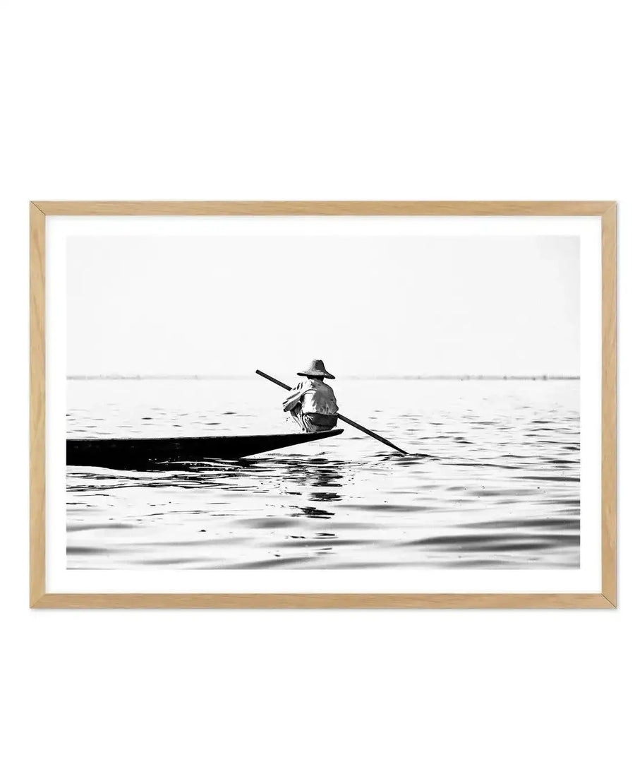 Framed black and white photograph of a person in a boat on calm water, with a natural wood frame and white border. The person, wearing a conical hat, is seen from behind, sitting low in a narrow boat and holding a long oar. The water shows subtle ripples and reflects the boat, with a faint horizon line in the distance under a bright sky.