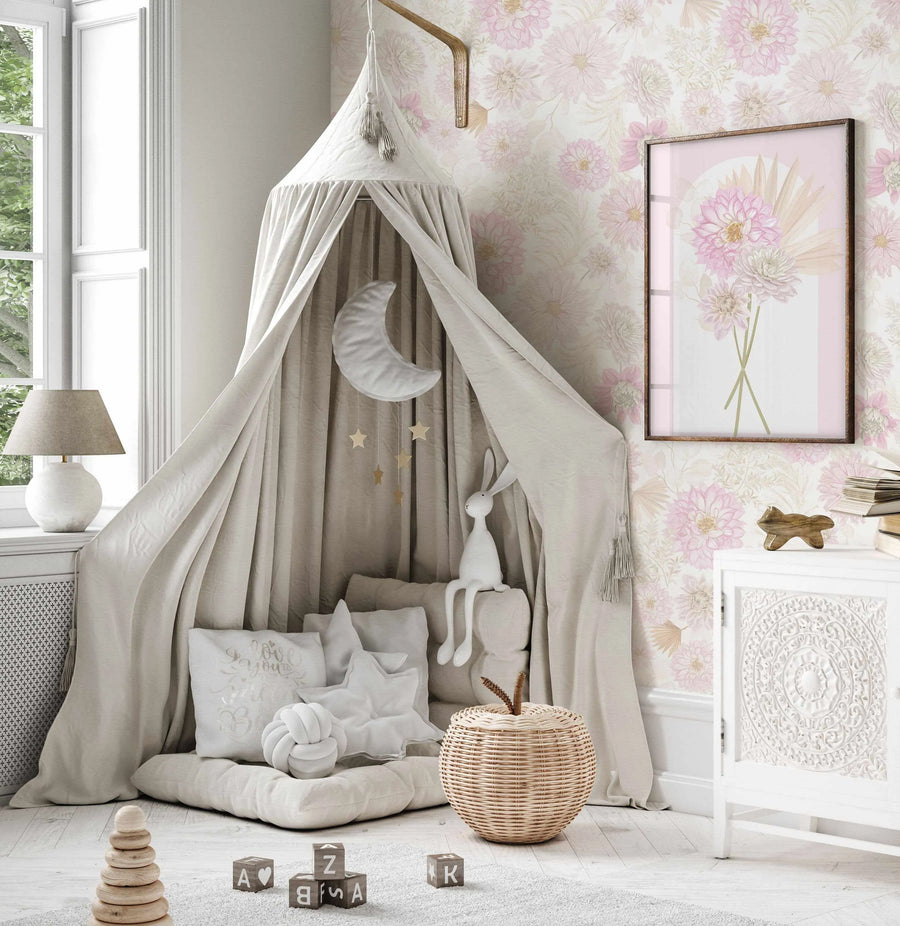 Lifestyle shot of a child's bedroom with a peel and stick wallpaper featuring a repeating pattern of hand-painted dahlias in soft blush pink and muted cream with delicate green foliage on a light cream background. The wallpaper covers the wall behind a white canopy tent with a plush moon and star mobile, a white rabbit toy, and soft pillows. A framed art print with a similar dahlia design hangs on the wallpapered wall.