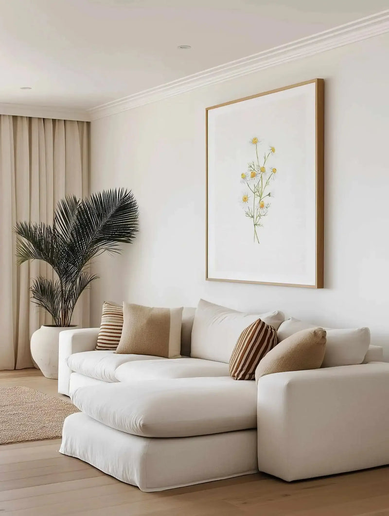 Framed art print displayed on a white wall above a curved cream-colored sofa. The artwork features a delicate watercolor illustration of yellow daisies with green stems arranged in a loose, naturalistic composition on white background. The print is set in a natural wood frame with white border. The room features neutral tones including beige curtains, a potted black palm plant, striped throw pillows in cream and tan, and a natural wood floor with woven area rug, creating a calm, minimalist interior setting.