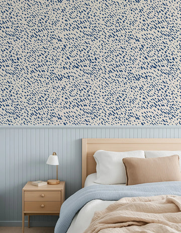 "Bedroom featuring Dapple Wallpaper with a repeating pattern of small, hand-painted dash marks in deep navy blue on a warm cream background, creating a textured, organic look. The wallpaper is displayed above light blue wainscoting and a natural wood bed frame with white, light blue, and beige bedding, and a wooden bedside table with a white lamp."