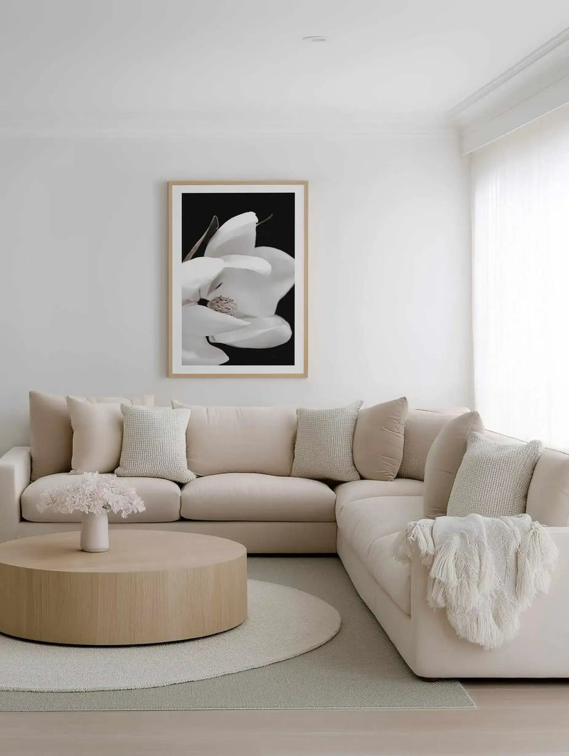 Black and white photographic art print of a magnolia flower displayed in a natural wood frame with white border, hanging above a curved cream sectional sofa in a minimalist living room with neutral tones, round wooden coffee table, and layered textures including linen pillows and a cream throw blanket.