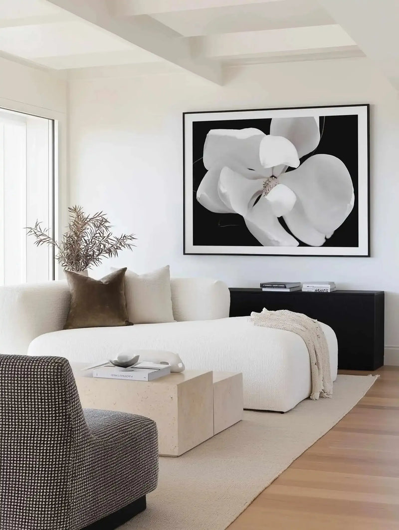 Dark Magnolia II art print displayed in a black frame with white border, hanging on a white wall above a black console table. The print features a close-up photograph of white magnolia petals with delicate stamens against a black background, creating striking contrast. The artwork is shown in a minimalist living room setting with a cream curved sofa, textured gray armchair, light wood coffee table, potted dried grasses, and warm wood flooring.