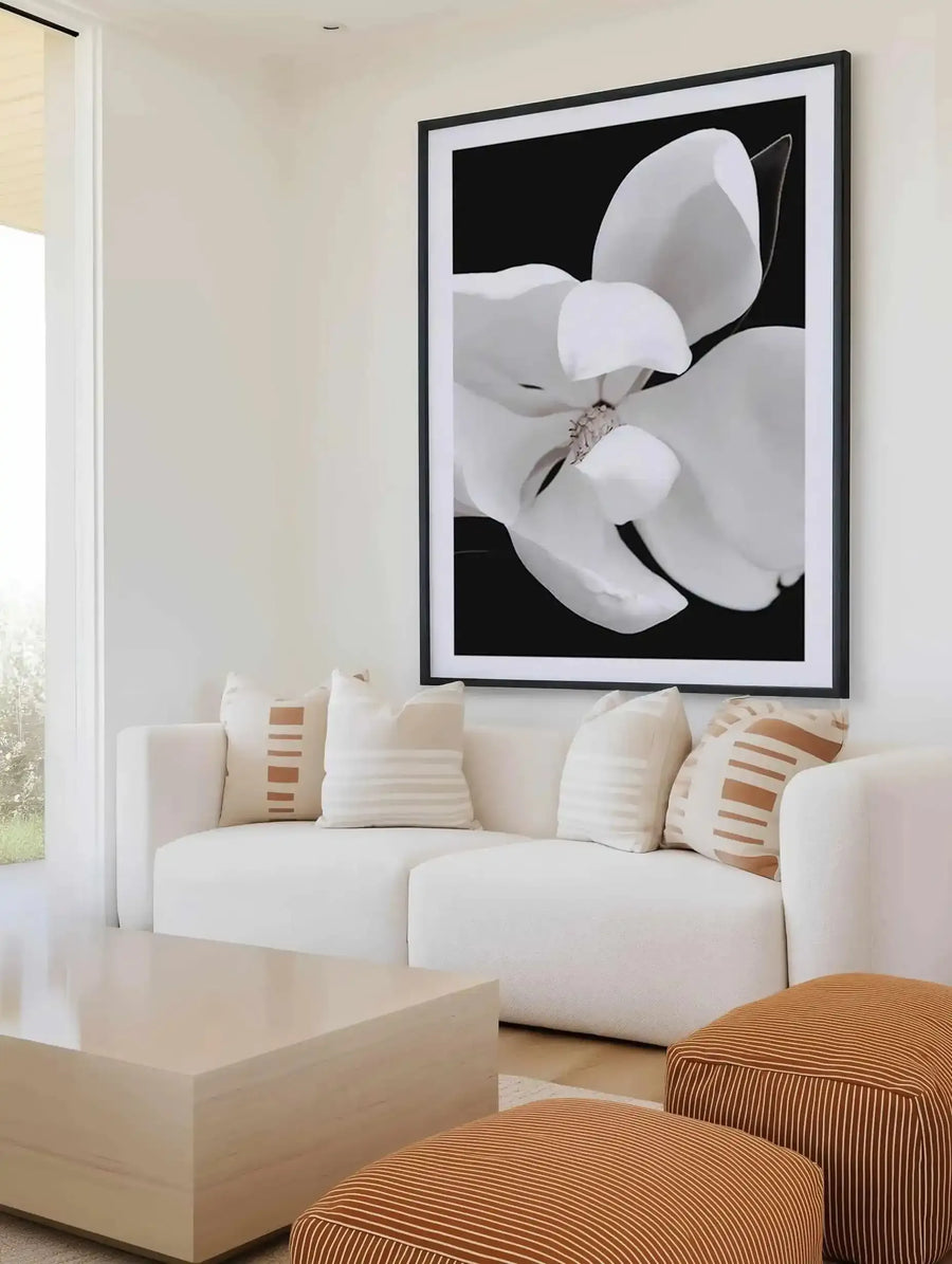 Dark Magnolia II art print displayed in a black frame with white border, hanging above a cream-colored sectional sofa. The artwork features a close-up botanical photograph of white magnolia petals against a black background, with soft gray tones and delicate details of the flower's center. The print is shown in a modern living room setting with natural light from a nearby window, cream cushions with tan and white striped accents, and a light wood coffee table in the foreground.