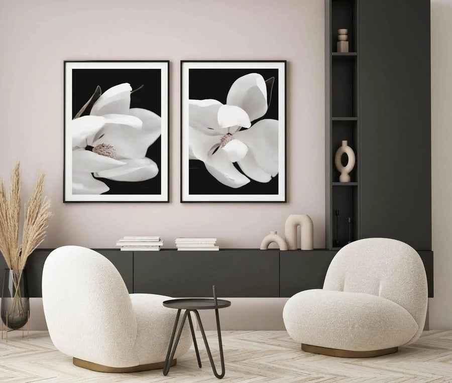 Framed art print displaying a close-up photograph of a white magnolia flower against a black background, shown in a black frame with white border. The left panel captures the flower's curved petals and visible stamens in soft focus, while the right panel shows another magnolia bloom from a different angle with layered petals. The diptych is displayed above a dark grey sideboard in a minimalist living room with cream-coloured rounded chairs, dried pampas grass, and ceramic decorative objects.