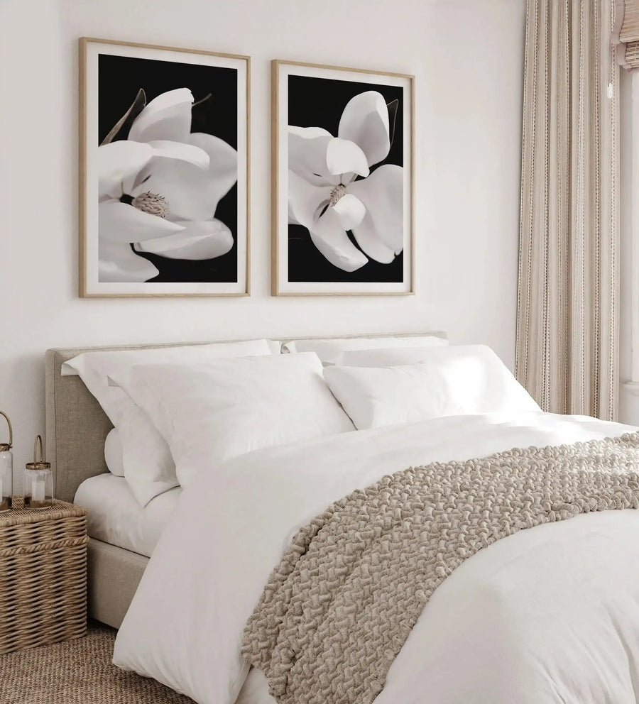 Dark Magnolia I art print displayed in a natural wood frame with white border, hanging on a cream wall above a beige upholstered bed. The print features a close-up photograph of a white magnolia flower with delicate, curved petals against a deep black background, showing fine detail of the flower's center stamens. The bedroom setting includes white bedding, a chunky knit throw blanket in warm beige, striped curtains, and a woven nightstand basket, creating a serene, minimalist aesthetic.