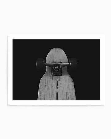 Framed black and white photograph of the underside of a longboard skateboard, centered against a dark background. The longboard deck is light-toned wood with visible grain and two dark, rectangular cutouts. A metal truck assembly with dark wheels is mounted near the top of the deck.