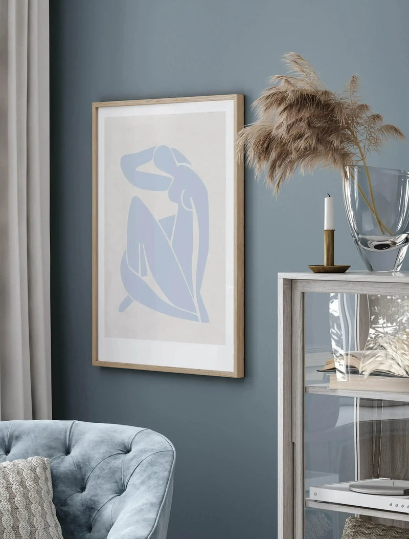 Lifestyle shot of a living room featuring a framed art print displayed on a dark blue wall above a light blue tufted sofa. The print features a minimalist abstract illustration of a reclining female figure in a light blue hue on a cream background, with a natural wood frame and white border. To the right of the print, a clear glass vase holds dried pampas grass, and a white candle in a brass holder sits on a light wood and glass display cabinet.