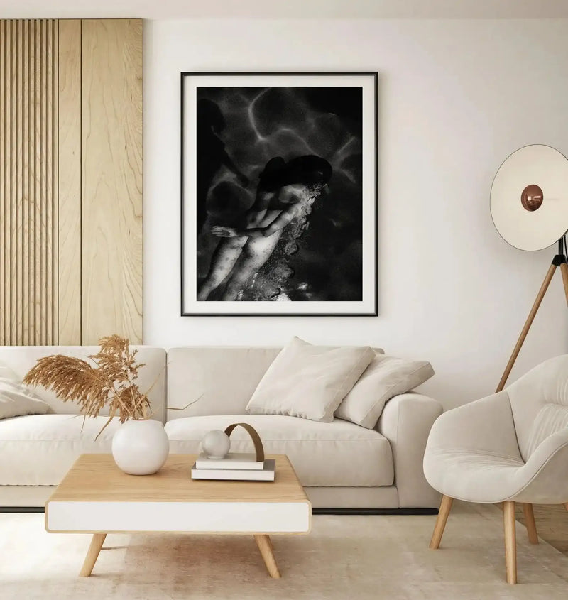 Black and white fine art photograph in black frame with white border, displayed on cream wall above cream upholstered sofa. The photograph shows a close-up portrait of a person with head tilted back, hands near face, rendered in dramatic chiaroscuro with high contrast lighting and ethereal smoke or mist effects in the background. Natural wood furniture and minimalist decor surround the framed print in a contemporary living room setting.