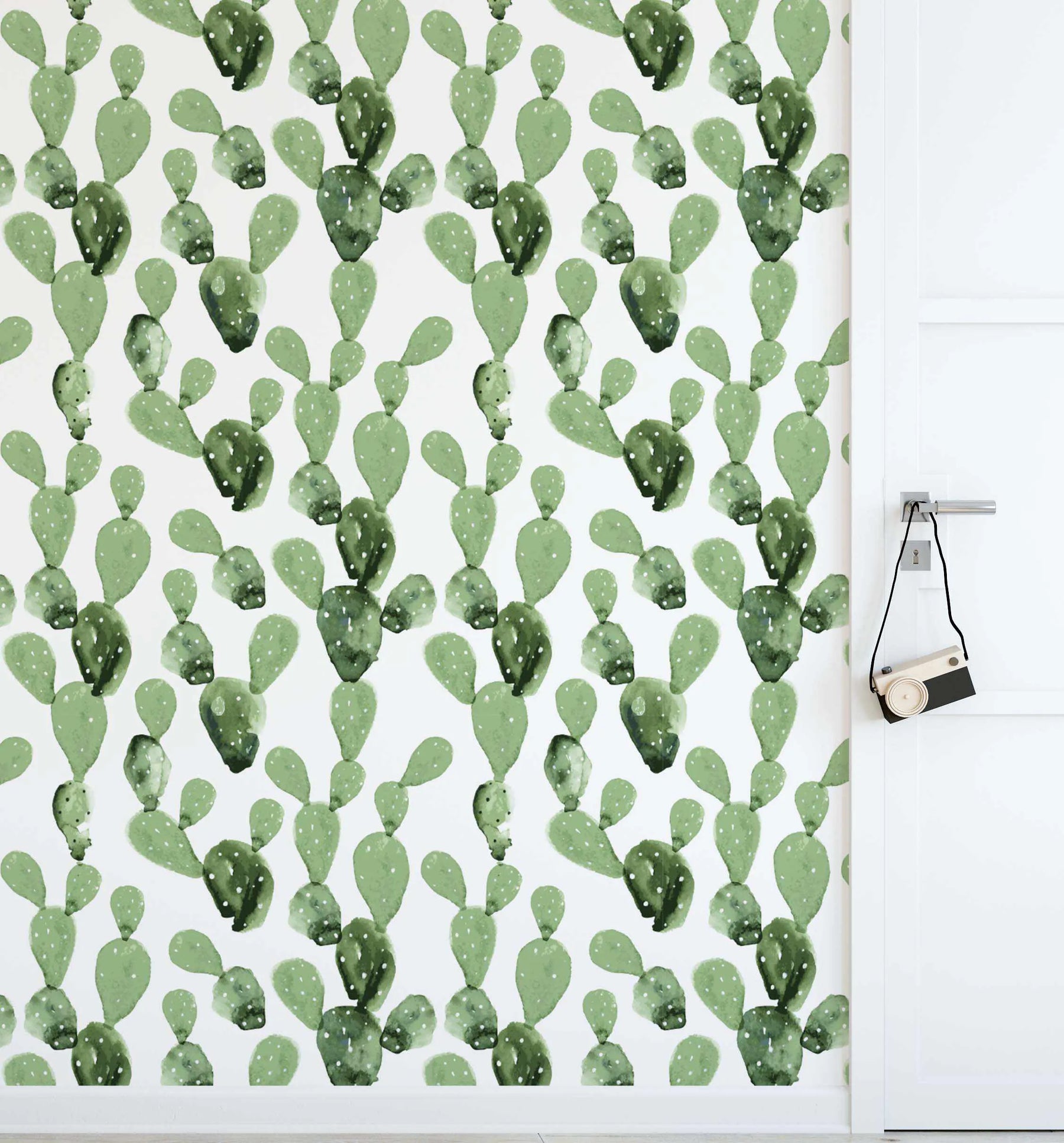 "Peel and stick wallpaper featuring a repeating pattern of hand-painted prickly pear cacti in various shades of sage green and deep olive on a soft white background. The botanical design has a loose, organic watercolour quality with visible brushstroke texture and small white dots representing spines. A white door with a silver handle and a small wooden toy camera hanging from it is visible on the right side of the image."
