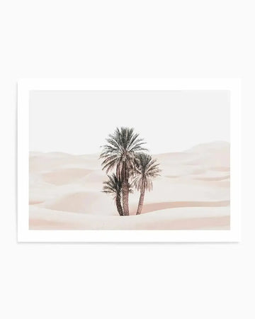 Framed photography art print featuring three palm trees in a vast desert landscape with soft, rolling sand dunes under a bright, clear sky. The sand has a pale, dusty rose hue, and the palm trees are rendered in muted greens and browns, creating a serene and minimalist composition. The print is displayed with a white border and a simple white frame.
