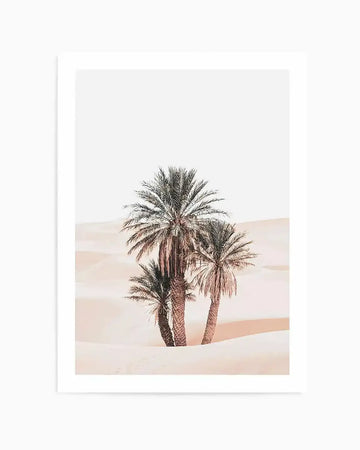 Framed photography art print featuring three palm trees in a desert landscape with soft, pale pink sand dunes and a bright white sky. The palm trees have dark green fronds and textured brown trunks, casting subtle shadows on the sand.