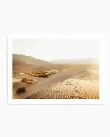 Framed photograph of a desert landscape at sunset with a natural wood frame and white border. The composition features rolling sand dunes with sparse green and yellow desert shrubs, and distant mountains under a bright, hazy sky with a warm sun flare on the right.