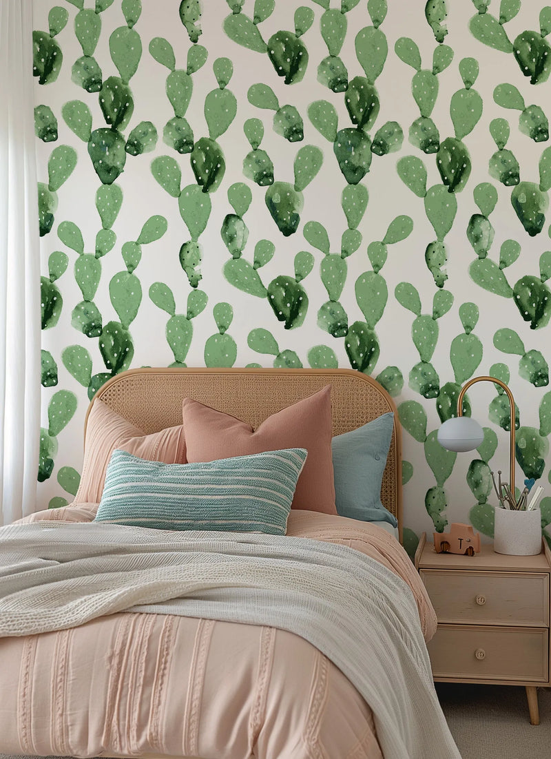 Lifestyle shot of a bedroom featuring peel and stick wallpaper with a repeating pattern of hand-painted prickly pear cacti in various shades of sage and emerald green with small white dots, on a soft white background. The wallpaper is visible behind a bed with a light brown rattan headboard, pink bedding, and pillows in dusty rose, light blue, and a striped teal and white pattern. A light-colored wooden nightstand with two drawers and a gold lamp with a light blue shade is to the right of the bed.