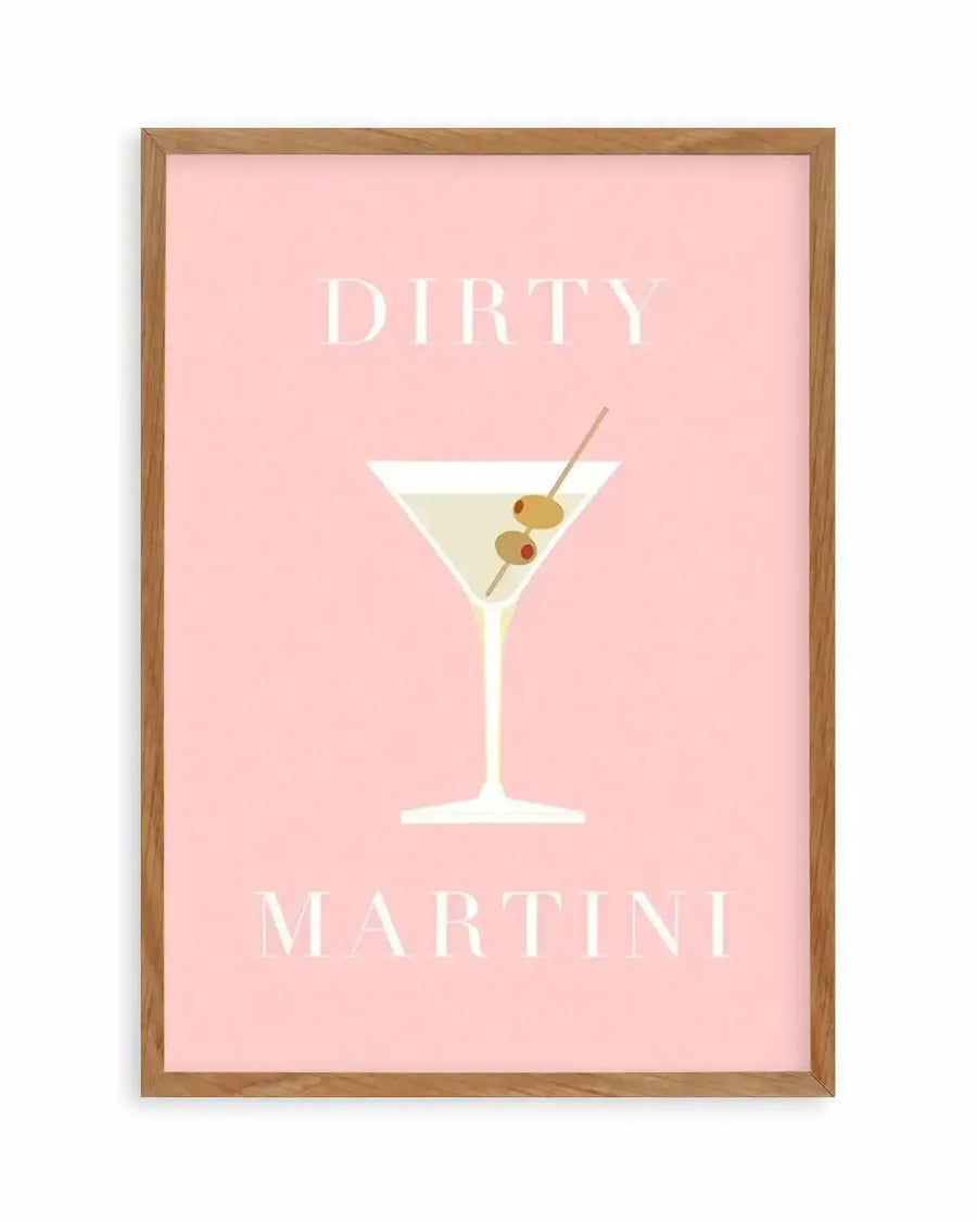 Art print featuring a minimalist illustration of a dirty martini cocktail on a solid light pink background, displayed in a natural oak frame with white border. The martini glass is white with a pale yellow liquid inside, garnished with two green olives and one red pimento on a toothpick. The words "DIRTY" and "MARTINI" are written in white serif font above and below the cocktail illustration.