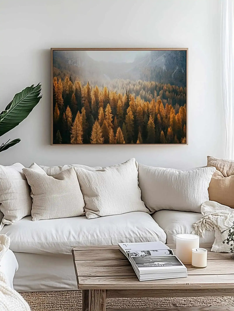 Lifestyle shot of a minimalist living room with a large framed canvas art print displayed above a white sectional sofa. The print features a photograph of a dense forest of golden-orange autumn trees in a valley, with misty mountains in the background, set in a natural wood frame. The sofa is adorned with white and cream textured pillows, and a wooden coffee table holds books and candles.