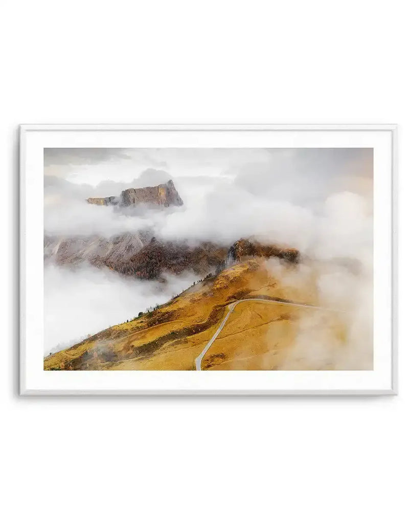 Framed art print of the Dolomites mountains in Italy, featuring a golden-yellow alpine meadow in the foreground with a winding gray mountain road. Dramatic rocky peaks rise through thick white clouds and mist in the background, with forested slopes visible beneath. The photograph captures a misty, atmospheric mountain landscape with warm ochre and brown earth tones contrasting against white clouds. The print is displayed in a thin silver frame with white border.