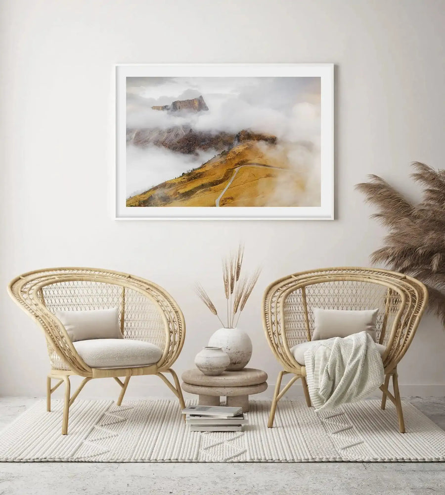 Framed landscape photograph of the Dolomite Mountains in Italy, featuring golden-yellow rolling hills in the foreground with a winding gray road, layered with misty white clouds obscuring rocky peaks in shades of brown and gray in the background. White wooden frame with white border. Displayed above two natural woven rattan chairs with cream cushions flanking a small round wooden side table with white ceramic vessels and pampas grass, on a cream and white striped area rug.