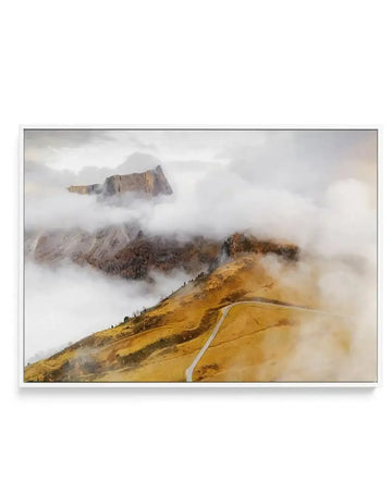 Framed canvas print of a misty mountain landscape in the Dolomites, Italy, with a white frame. The composition features a winding road on a golden-brown hillside in the foreground, partially obscured by soft, white clouds. In the midground, dark green trees are visible through the mist, leading to rugged, grey-brown mountains in the background, also partially enveloped in clouds. The overall mood is serene and atmospheric.