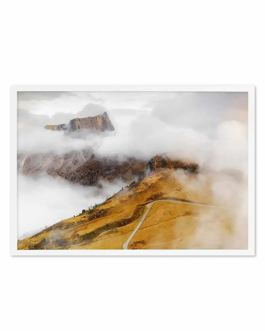 Framed art print of the Dolomites Mountains in Italy, featuring a golden-yellow mountainside in the foreground with a winding road cutting through it. Misty white clouds obscure the middle and background layers of jagged peaks and forested slopes in shades of brown, gray, and muted green. The sky is overcast and pale. The photograph has a soft, atmospheric quality with layered depth created by the fog. White frame with white border.
