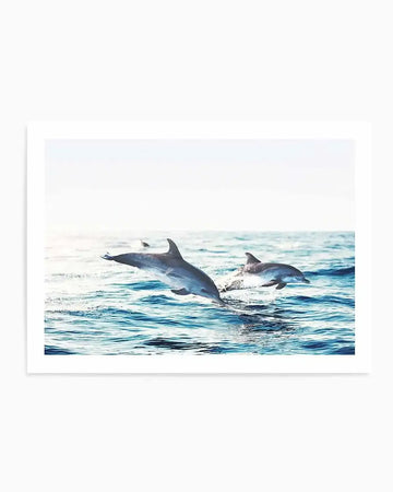 "Art print featuring a vibrant photograph of two dolphins leaping out of the ocean. The larger dolphin on the left is mid-air, while the smaller one on the right is just breaking the surface, creating a splash. The water is a deep blue with visible ripples and white foam, under a bright, clear sky."