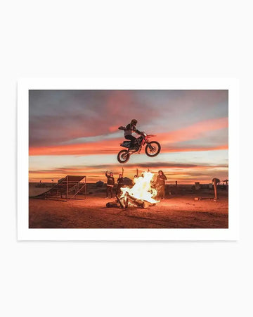 "Art print featuring a motocross rider mid-air over a bonfire at sunset, with a vibrant sky displaying hues of orange, pink, and purple. The rider is silhouetted against the colorful sky, with spectators gathered around the bonfire below, and a dirt ramp visible on the left."