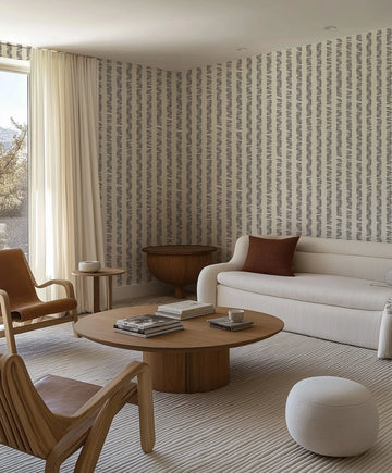 Dots n Dash Stripe Wallpaper displayed on a living room wall in a neutral beige and grey colorway. The design features vertical stripes composed of small repeated dots and dashes in a woven, textured pattern. The wallpaper covers the entire back wall behind a cream sofa with brown cushion, wooden accent chairs, and a round wooden coffee table. Natural light streams in from a floor-to-ceiling window with cream curtains, highlighting the subtle texture and neutral tones of the wallpaper design.