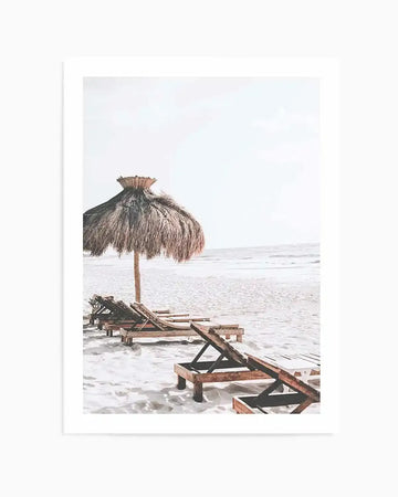 Framed photography art print of a serene beach scene with a thatched umbrella and several wooden lounge chairs on white sand, extending towards the gentle waves of the ocean under a bright, overcast sky.