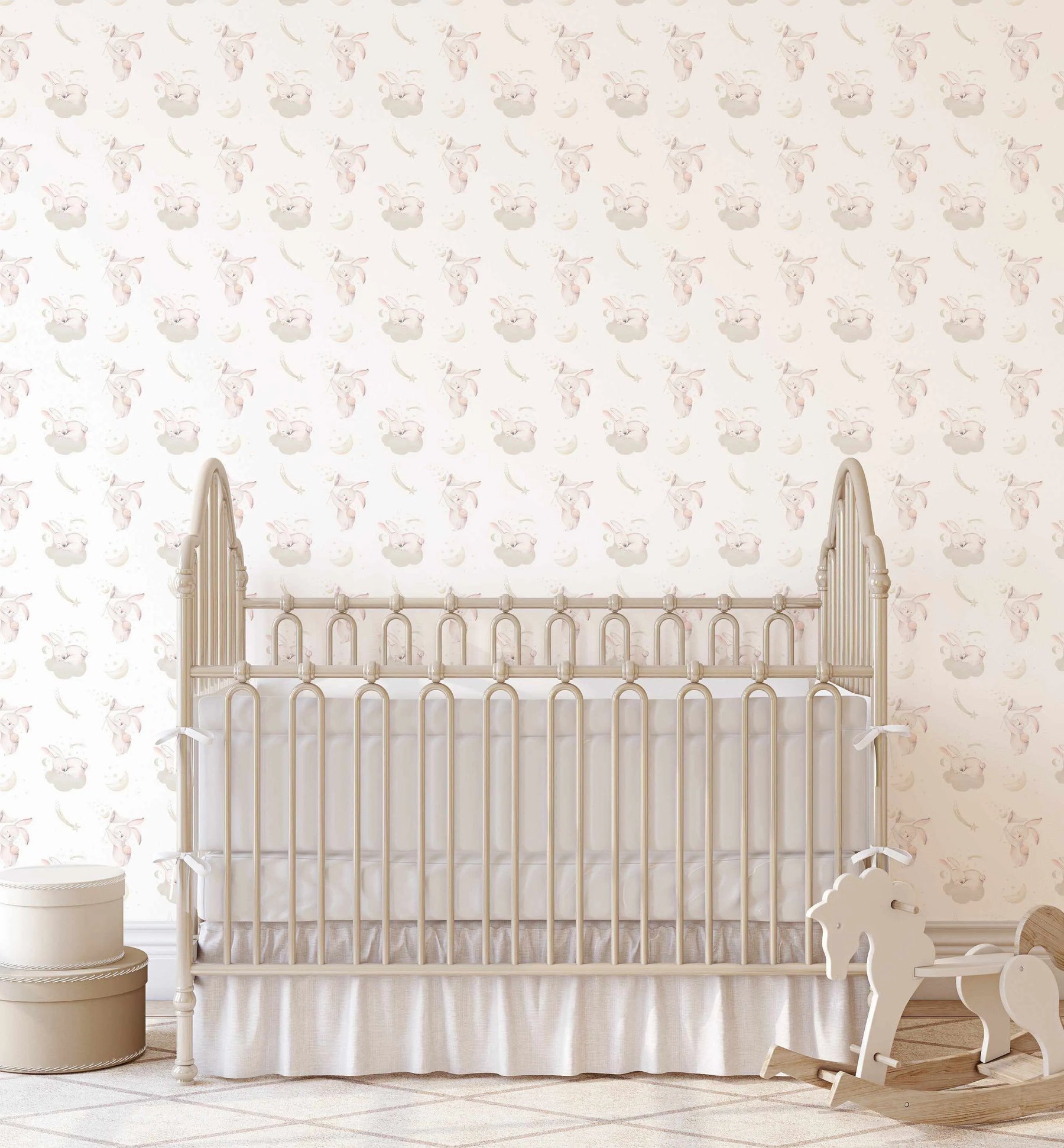 Lifestyle shot of a nursery with a crib and a rocking horse, featuring a wallpaper with a repeating pattern of sleeping bunnies on clouds, bunnies holding stars, crescent moons, and shooting stars in soft pink, grey, and cream tones on a light cream background. The wallpaper has a gentle, hand-painted watercolor quality.