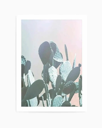 Dreamy Cactus II art print featuring a cluster of prickly pear cacti with flat, oval-shaped pads in shades of sage green and pale mint, silhouetted against a soft, dreamy sky in blush pink, pale blue, and cream. The photograph has a hazy, ethereal quality with diffused lighting. The print is mounted on a white background within a white frame.
