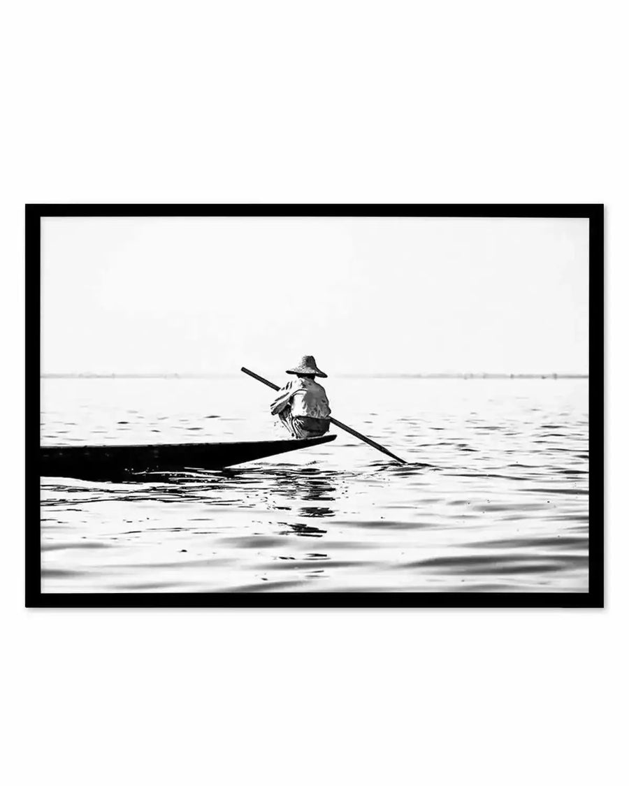 Framed black and white photograph of a lone person in a conical hat rowing a small boat on calm water, with a black frame and white border. The person is seen from behind, crouched in the boat, holding a long oar that extends into the water. The horizon is a soft, hazy line under a bright sky.