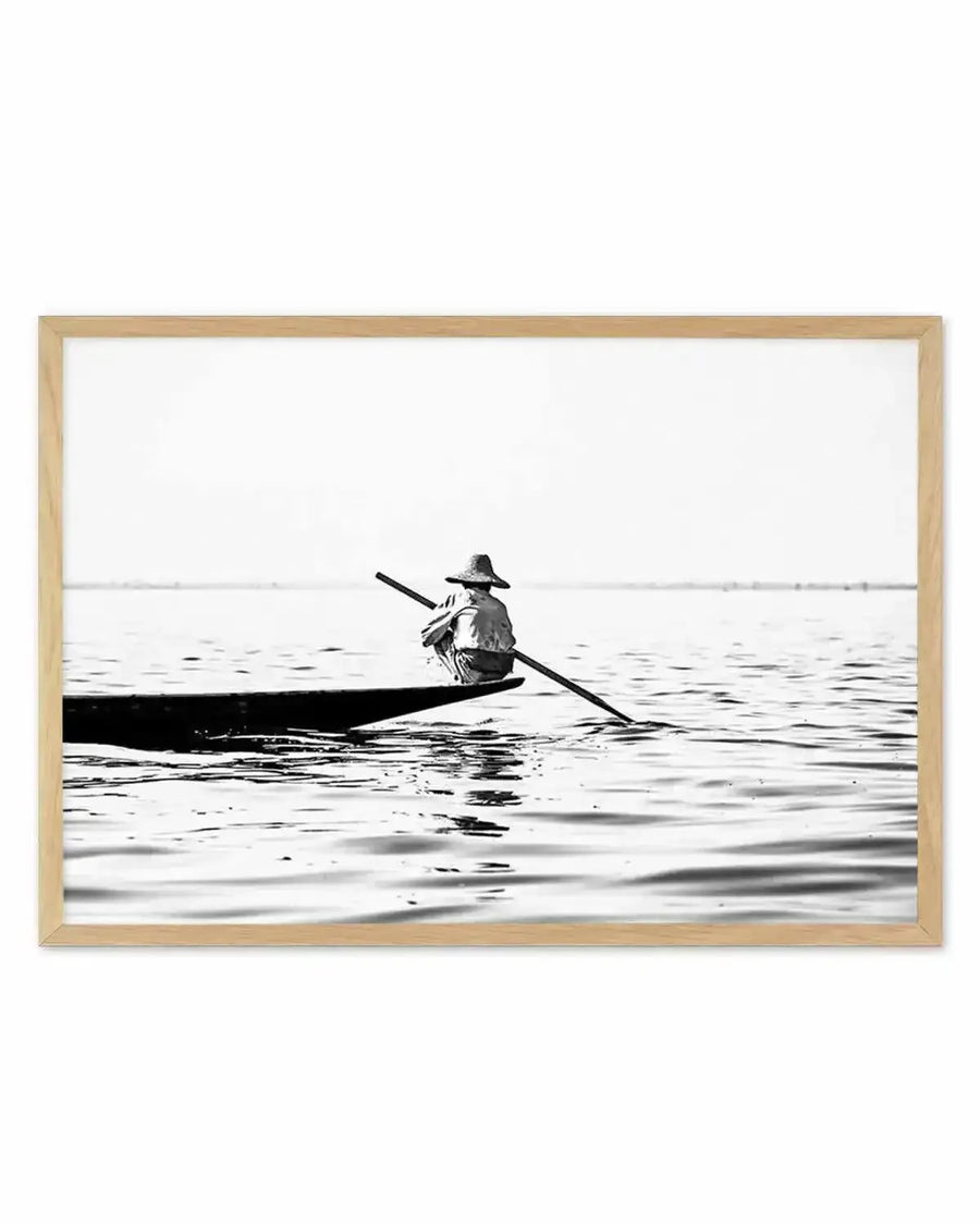 Framed black and white photograph of a lone person in a small boat on calm water, with a natural wood frame and white border. The person is wearing a conical hat and is positioned towards the back of the boat, holding a long oar that extends into the water. The composition features a vast, bright sky above the water, creating a serene and minimalist scene.