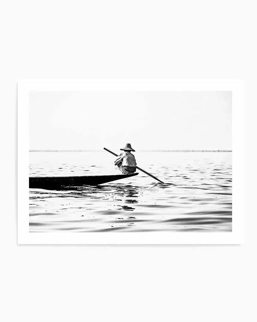 Framed black and white photograph of a lone person in a small boat on calm water, with a natural wood frame and white border. The person, wearing a wide-brimmed hat, is seen from behind, paddling with a long oar. The composition features a wide expanse of water with subtle ripples and a bright, clear sky above.