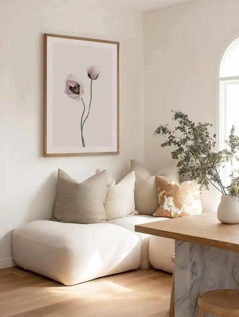 Dining Nook:
Large framed art print displayed above a curved cream sofa in a minimalist dining nook. The print features two delicate poppy flowers with soft pink and purple petals and slender dark stems against a warm cream background, with a natural wood frame and white border. The room includes a light wood dining table with a marble base, a white vase with dried branches, and light wood flooring.