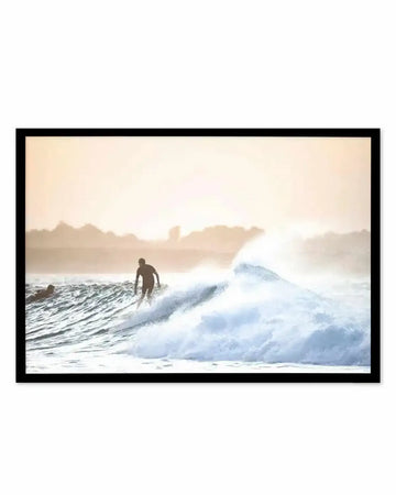 Framed photograph of a surfer riding a large wave at dusk, with a black frame and white border. The composition captures the surfer silhouetted against the warm, hazy sky, with another surfer partially visible in the foreground. The wave is breaking with white foam and spray, and distant hills are visible on the horizon.