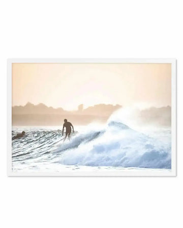 Framed photography art print featuring a surfer riding a large wave at dusk, with a white frame and white border. The composition captures the silhouette of the surfer against a warm, hazy sky, with another surfer visible in the background.