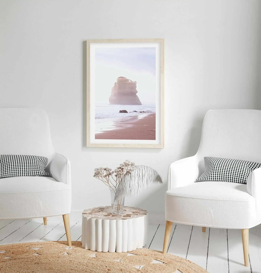Lifestyle shot of a minimalist living room featuring a framed art print of a coastal scene displayed above a round wooden side table. The print depicts a large rock formation on a sandy beach with gentle waves under a hazy, soft pink sky, framed in natural wood with a white border. Two white upholstered armchairs with houndstooth throw pillows flank the table, and a round jute rug covers the white wooden floorboards.